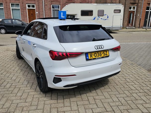 Zicht op een witte Audi met lesauto bord, geparkeerd op een straat.